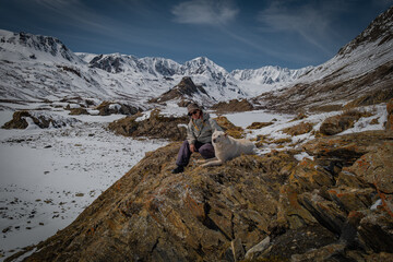 Naklejka premium Female traveler sitting with white dog on rocky hill, woman hiker resting with pet on scenic snow mountains background, winter adventure and friendship lifestyle, trekking, serenity