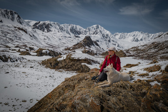 Female traveler sitting with white dog on rocky hill, woman hiker resting with pet on scenic snow mountains background, winter adventure and friendship lifestyle, trekking, serenity