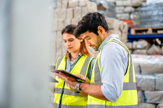 Diverse team of warehouse supervisors in safety vests using digital tablets to manage inventory and logistics in a large industrial printing facility storage area