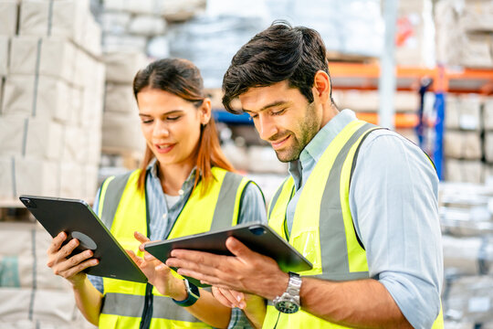 Diverse team of warehouse supervisors in safety vests using digital tablets to manage inventory and logistics in a large industrial printing facility storage area
