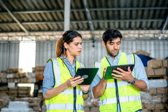 Diverse team of warehouse supervisors in safety vests using digital tablets to manage inventory and logistics in a large industrial printing facility storage area