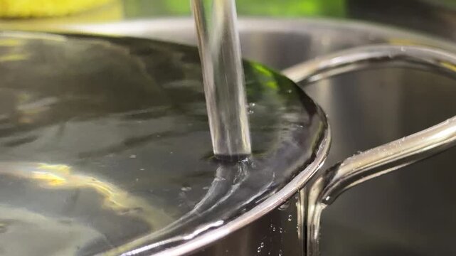 A stream of water flows from a kitchen tap into a stainless steel pot already filled to the brim. The continuous impact of the falling water against the surface creates a steady, resonant hum.