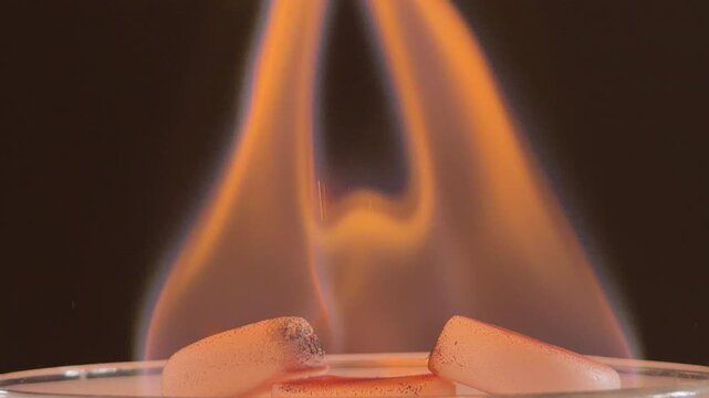 Solid white fuel tablets burn in a glass container. The intense orange flames rise upward, producing a quiet crackling and popping sound. This chemical combustion releases steady heat and light.