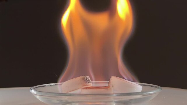 Three white fuel tablets burn steadily in a glass dish. The rising orange flames create a gentle crackling and popping sound as the material consumes. The fire glows brightly against dark background.