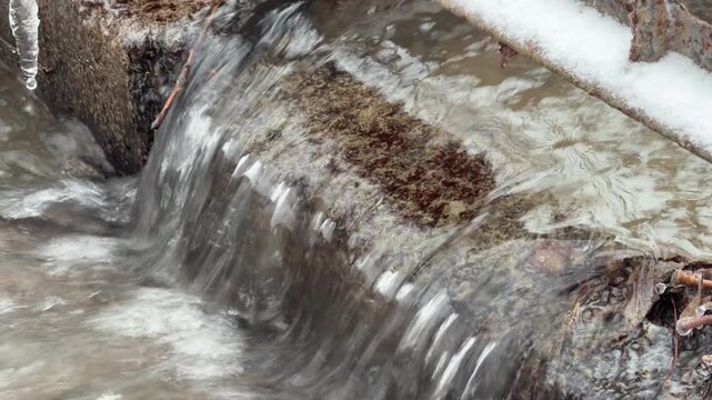 Rushing water flows over a low stone ledge into a wider stream. The movement of the heavy current produces a constant, loud churning and splashing sound. Small icicles hang nearby