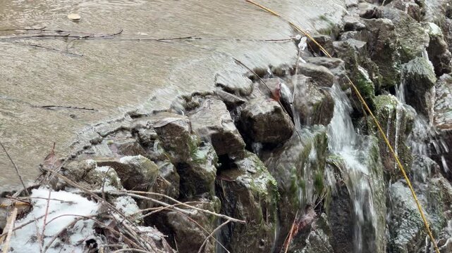 A stream of water cascades over a row of mossy rocks and icy debris. This constant movement generates a continuous splashing sound as the liquid hits the stones below.