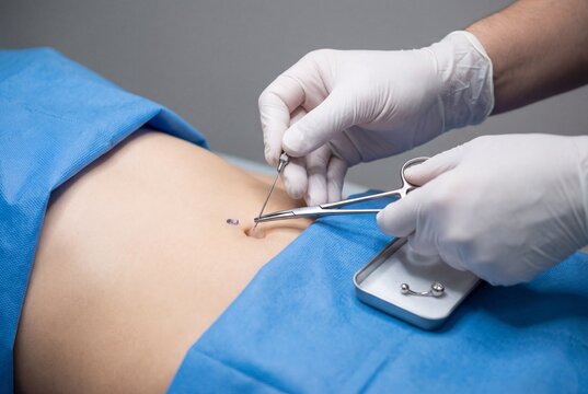 Professional piercer in white gloves performing belly button piercing on female abdomen with needle
