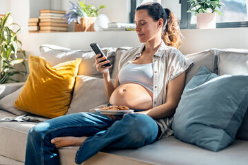 Obraz premium Pregnant woman using smartphone while eating a cookies sitting on the couch at home