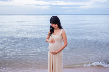 Pregnant woman standing on a beach with beautiful evening sea holding baby with love and care.