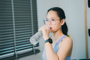 Young woman drinking water clear plastic bottle indoor casual wear fitness tracker eyeglasses ponytail hairstyle hydration healthy lifestyle