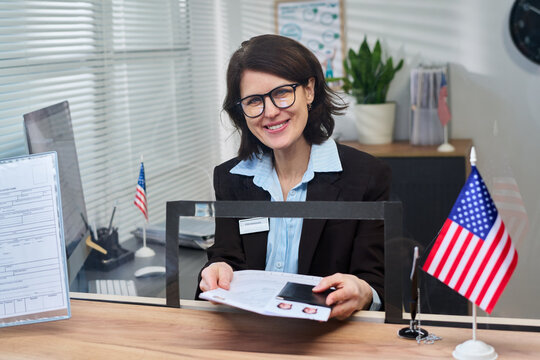 Portrait of middle aged woman sitting at visa center desk smiling while holding documents and passport photos, American flags and paperwork visible on counter, office background