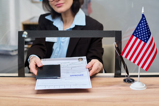 Caucasian middle aged woman handing approved visa application form and passport through service window at visa center, American flag standing on counter, partial face visible