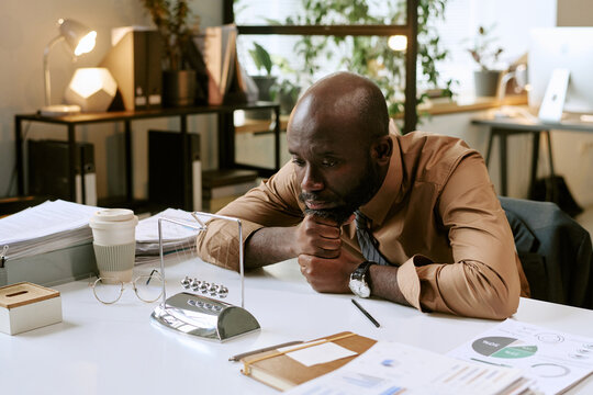 Black man sitting at desk leaning forward watching Newtons cradle, procrastinating in modern office workspace, business documents and coffee cup on table