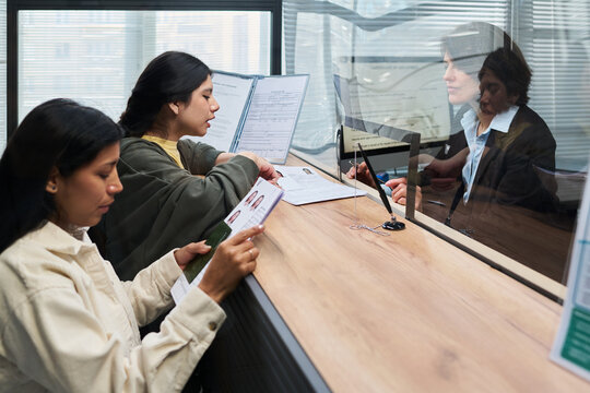 Two young adult Hispanic women submitting documents at visa center counter, interacting with middle aged Caucasian woman behind glass partition, holding identification papers