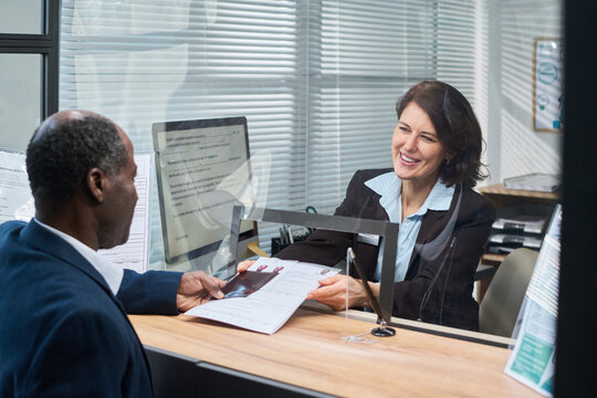Middle aged Caucasian woman smiling while assisting middle aged Black man submitting documents at visa center reception desk, both engaging in formal application process through glass partition
