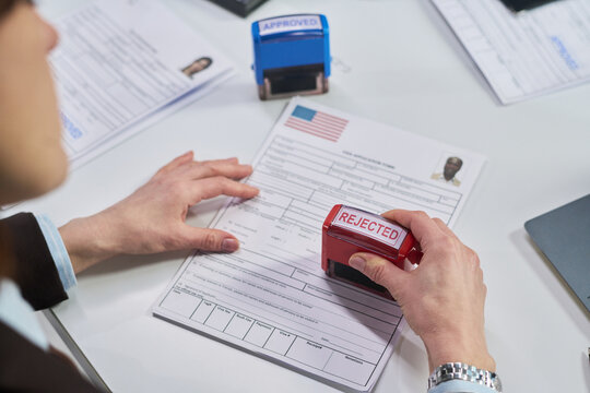 Caucasian middle aged woman stamping visa application form with rejected stamp at visa center, visible hands holding document, official paperwork and approval stamp on desk