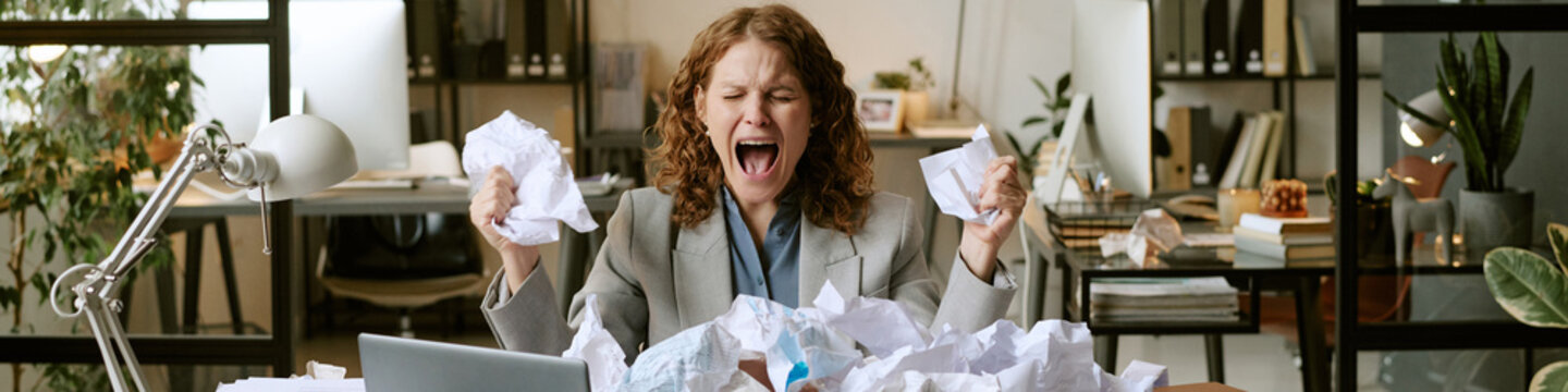 Caucasian young adult woman sitting at desk screaming while holding crumpled papers, surrounded by messy piles of documents, expressing frustration in modern business office