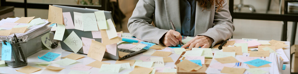 Caucasian woman working at cluttered desk covered with sticky notes, writing on paper beside open laptop, demonstrating busy business planning and organization process © AnnaStills