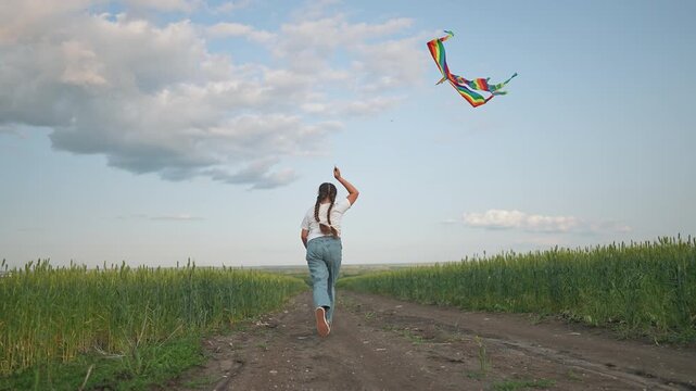 The girl runs in the summer field, holding her kite high. The wind lifts the kite into blue sky. She loves to play in the outdoor sun. The summer field, sky, wind, and kite bring endless fun for girl.
