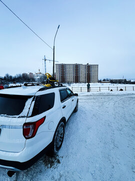 Winter street scene with a white Ford Explorer SUV and ski rack near apartment buildings and a snowy park