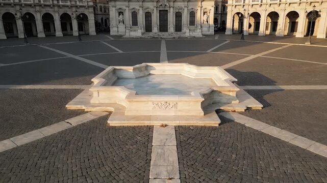 A white marble fountain sits in a large plaza, surrounded by classical architecture