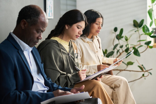 Middle aged Black man and two Hispanic girls sitting in visa center waiting area filling out paperwork, focused on completing application forms