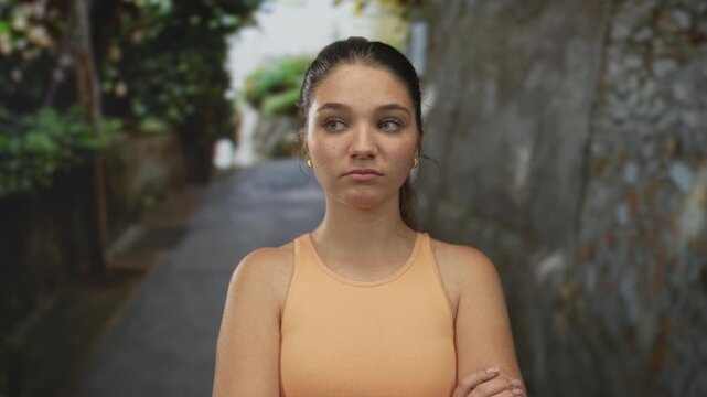 Caucasian teenage girl with bare arms crossed glancing upward in a narrow stone street framed by walls; rejection.