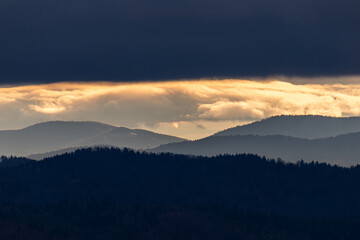 Obraz premium Layers of misty mountain ridges in Beskidy, Poland, under a dramatic dark sky with a glowing golden sunset light stripe.
