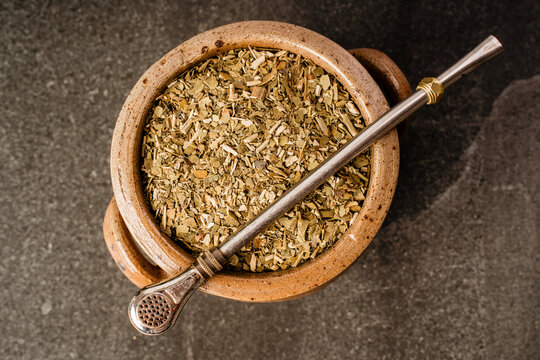 Dried yerba mate leaves in a rustic ceramic vessel with metal bombilla placed on a dark textured background in a simple still life composition.
