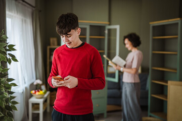 Teenage boy using smart phone while mother reading book at home