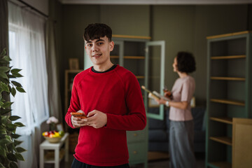 Teenage boy using smart phone while mother reading book at home