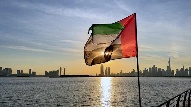 United Arab Emirates flag fluttering warm sunset light with city skyline silhouette and waterfront. National celebration, tourism landmark, Arabian Gulf coast, cultural heritage and progress concept
