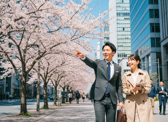 サクラピンクの並木道で談笑する、ベージュのトレンチコートを纏った女性とチャコールグレーのスーツを着た男性の日本人新社会人ビジネスカップル