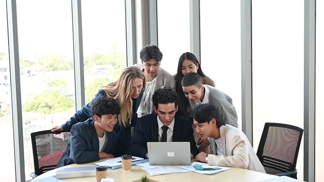 Business team office meeting collaboration around laptop with coworker and young adult diverse group leaning over computer planning project in bright modern office, focused teamwork brainstorming buzz