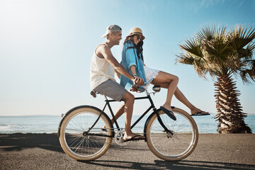 Fototapeta premium Happy, couple and travel on bike at beach for holiday adventure, sightseeing nature or bonding. Support, people and rental bicycle in road for vacation ride, healthy relationship and space in summer