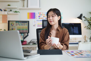 Graphic designer holding a coffee cup while working on digital tablet and laptop at her desk. Creative lifestyle and flexible workspace concept.