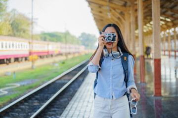 Fototapeta premium A girl smiles, holding a film camera. Young Asian woman traveler with a camera taking pictures on a train platform. Adventure, hangout, travel, holiday, relaxation, photography concept.