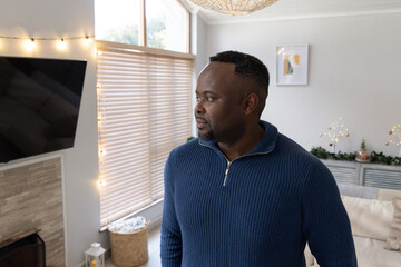 Naklejka premium African American man standing by window in living room wearing navy sweater, viewing string lights
