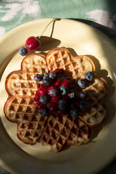 Top view close-up of a round waffle with blueberries, cherries, and jam on a yellow plate