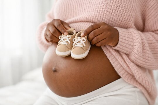 Close up of pregnant African American woman holding small baby shoes on her bare belly, unrecognizable future mother in pink sweater preparing for childbirth.