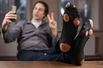 A man with holey socks sits on a chair and takes a selfie on his smartphone