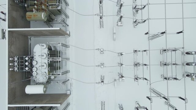 Gliding overhead view shows frosted oil filled transformers, busbars, and connections within an active winter switchyard.