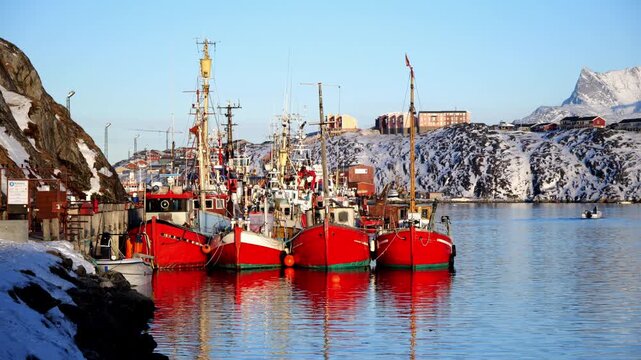 A line of iconic red wooden fishing vessels moored at a pier in Nuuk, Greenland. The background features colorful arctic houses and snow-covered mountains under a clear sky.