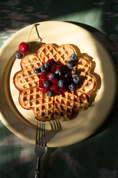 Top view close-up of a round waffle with blueberries, cherries, and jam on a yellow plate and fork