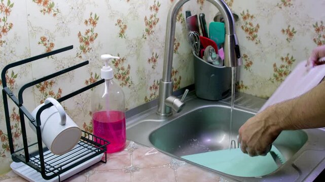 Hands are busy washing dishes in a sink. Items are rinsed and cleaned with a sponge and soap. The kitchen has a simple design with basic items nearby.