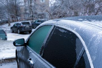 Close-up of a parked car with windows and roof coated in thick ice, in a residential area on a cold winter day.