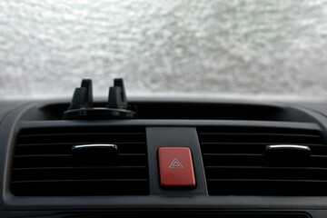 Close-up of a car dashboard showing the red emergency hazard button, air vents, and phone holder against a frosted window background.
