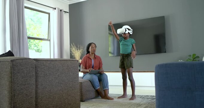 African American mother-daughter at home daughter putting on white VR playing mother tapping tablet