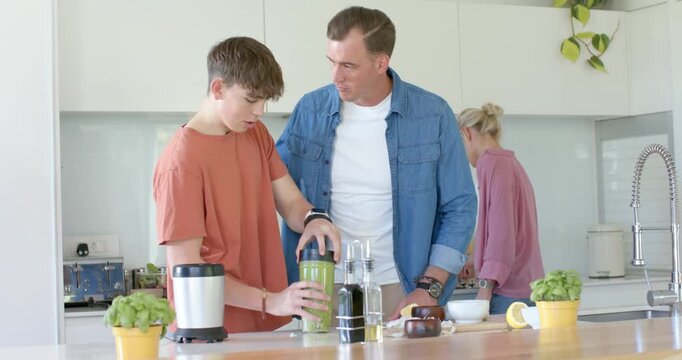 Father guiding teen son holding blender cup sealing lid checking and tasting smoothie in kitchen