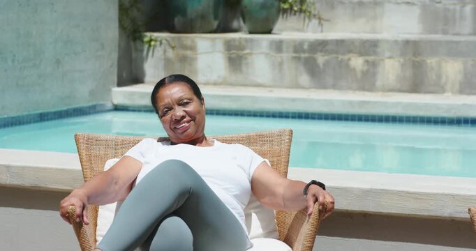 Senior African American woman sitting wicker seat by pool, shifting legs, smiling, posing at camera